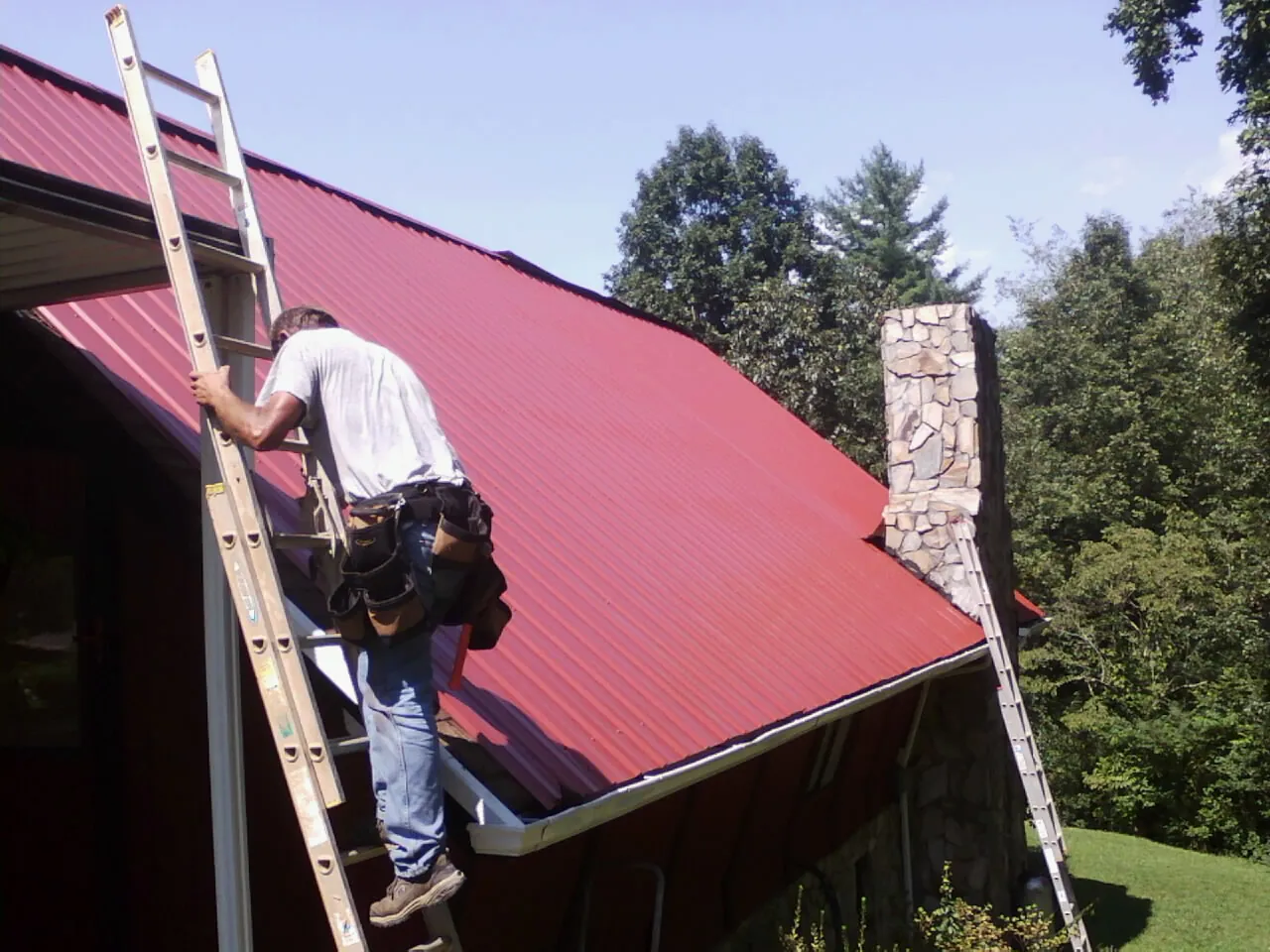 Roofer on a ladder beside a red metal roof and stone chimney — Soffit & Fascia Repair by Roofing Brothers in Kilbourne